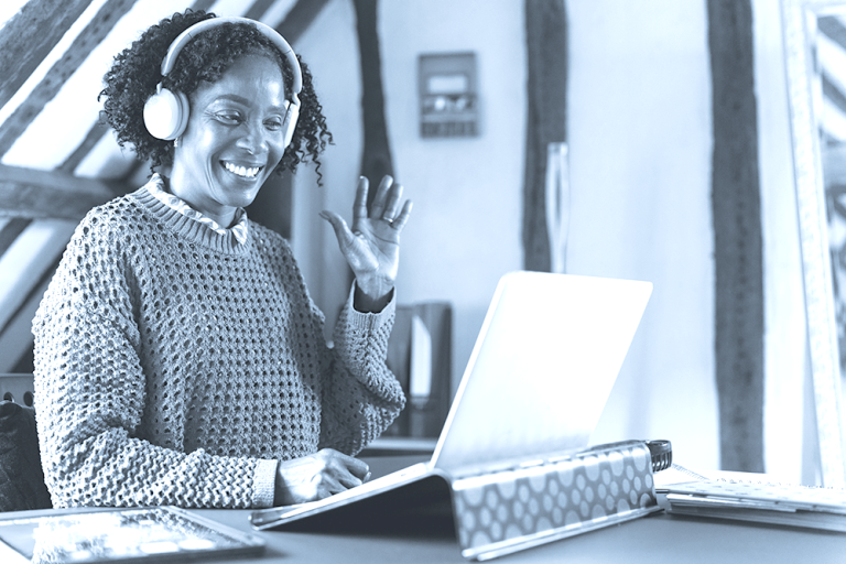 Woman wearing headphones smiles and waves at her laptop during a video call at her desk.