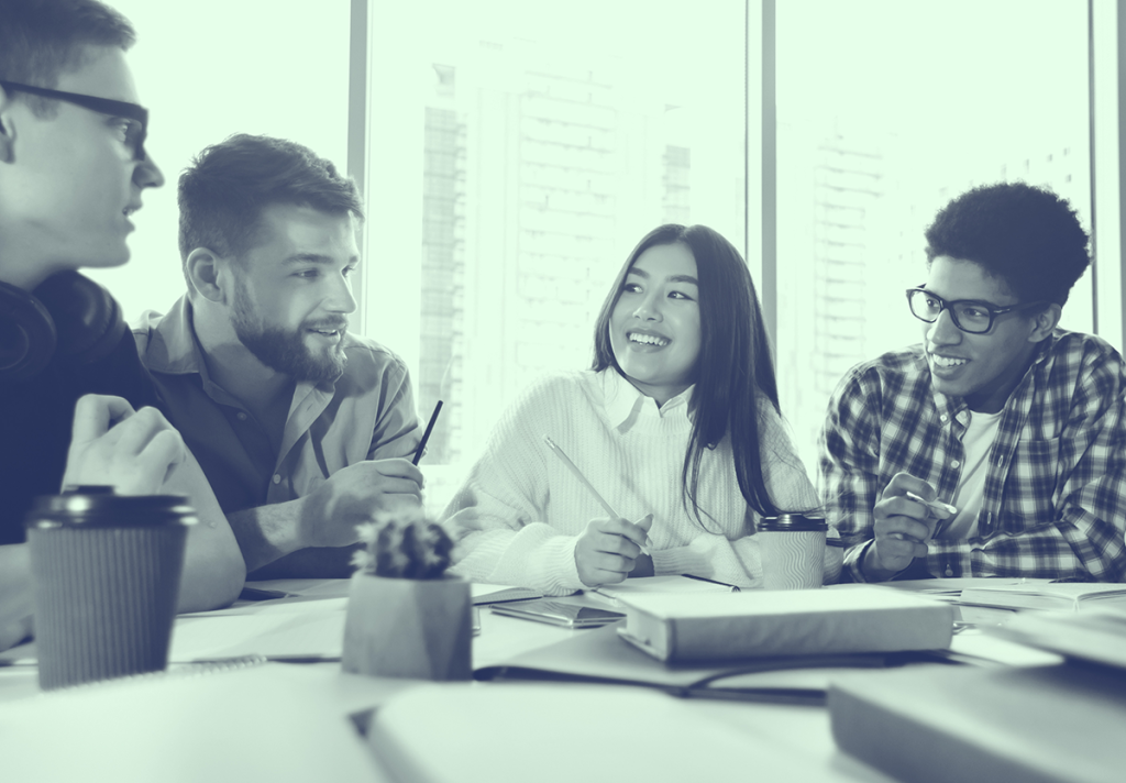 Four young adults sit at a table, talking and smiling, with notebooks, coffee, and papers in front of them.