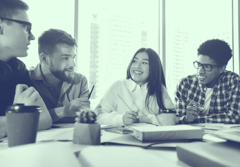 Four young adults sit at a table, talking and smiling, with notebooks, coffee, and papers in front of them.